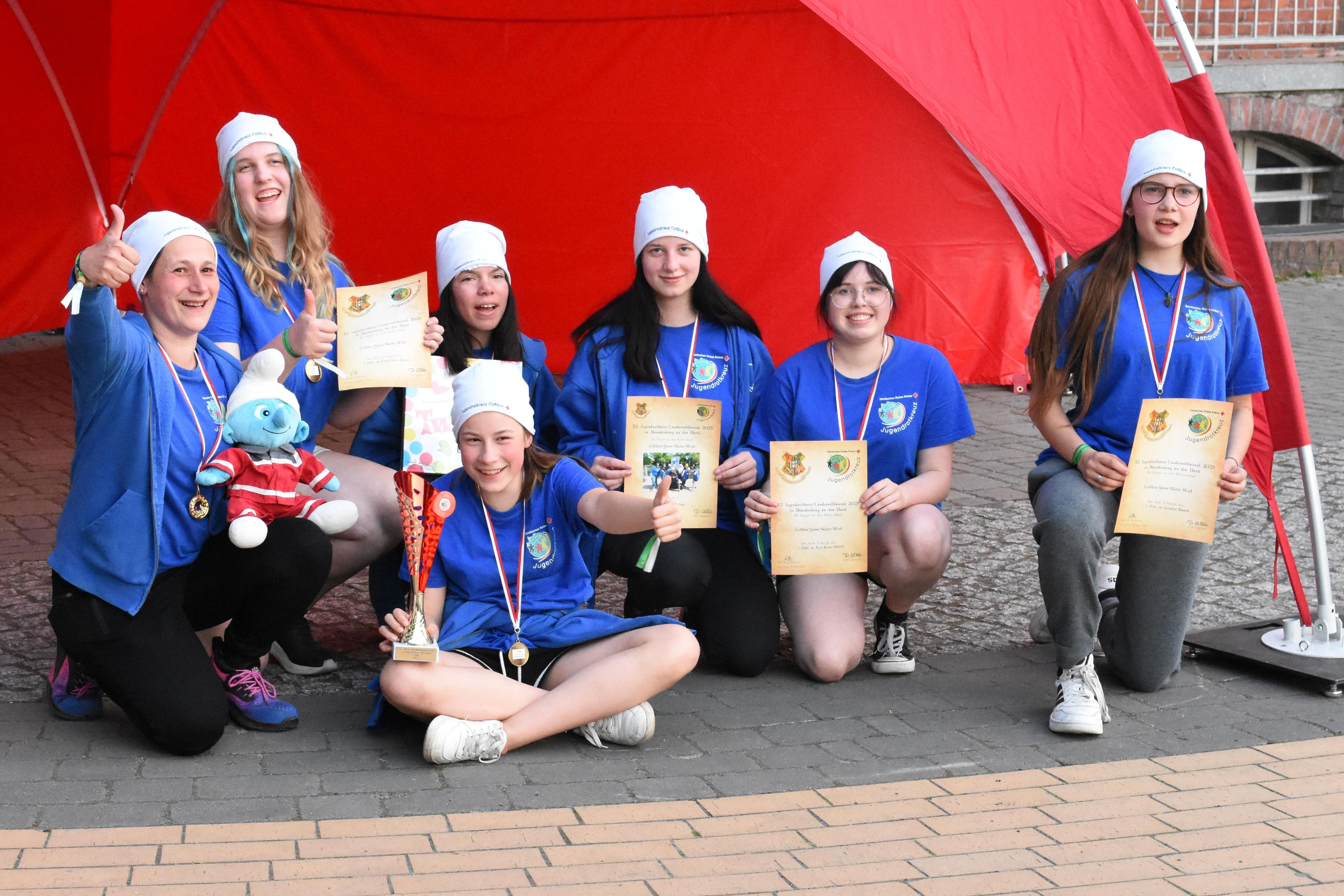 Eine Gruppe aus sechs Jugendlichen in blauer Kleidung mit Logo des Jugendrotkreuz sowie die Gruppenleiterin hocken und sitzen vor einem roten Pavillion. Sie lachen und halten teilweise die Daumen nach oben. Sie tragen Medallien und zeigen Urkunden nach vorn. Ein Mädchen hält einen Pokal.